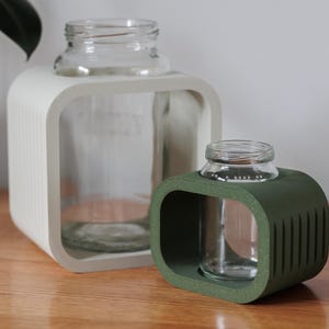Può includere: Due portavasetti decorativi in vetro. Uno è bianco, l'altro verde. Ogni supporto ha una forma quadrata con angoli arrotondati e un vasetto di vetro all'interno. Il supporto bianco è più grande di quello verde. I vasetti sono trasparenti.