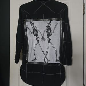May include: Black long-sleeve shirt with a white and gray graphic of two skeletons holding hands. The shirt has a silver zipper detail around the graphic and a curved hemline. The back of the shirt has white stitching.