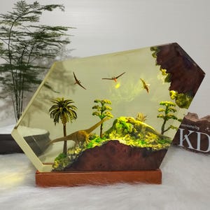 May include: A decorative resin lamp featuring a prehistoric scene with a long-necked dinosaur, palm trees, and flying pterosaurs. The lamp has a wooden base and a geometric shape, with a warm, yellow glow.