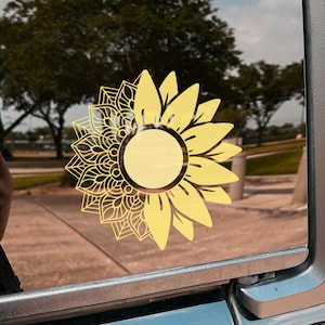 May include: A yellow sunflower decal with a mandala design on the left side is affixed to a car window. The sunflower has a circular center and pointed petals. The background shows trees and a blurred outdoor scene.