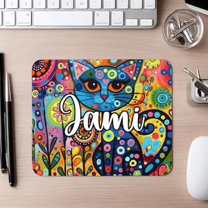 May include: A colorful mousepad featuring a stylized blue cat with orange eyes and a whimsical design. The mousepad has the name "Jami" in white script. The background is a vibrant mix of colors and patterns.