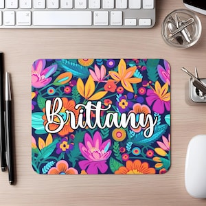 May include: A rectangular mousepad with a vibrant floral design in shades of pink, orange, yellow, and teal on a dark blue background. The name "Brittany" is written in white cursive across the center.