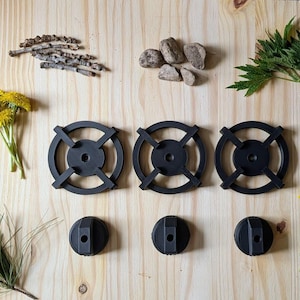 May include: An overhead shot displays three black metal burner grates and three matching knobs, arranged with natural elements like grass, yellow flowers, and stones on a light wood surface. The grates have a circular design with four spokes.