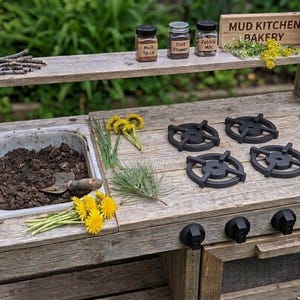 May include: A rustic wooden mud kitchen with a sink, stove burners, and oven knobs. It features jars labeled "Mud Spice," "Dirt Flower," and "Pebble Mix." The sign reads "Mud Kitchen Bakery." Dandelions and twigs are arranged on the surface.