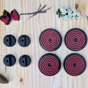 May include: An overhead shot of black and red spiral-patterned coasters and black, round objects with a rectangular hole. The items are arranged on a light wood surface with natural elements like leaves, flowers, and stones.
