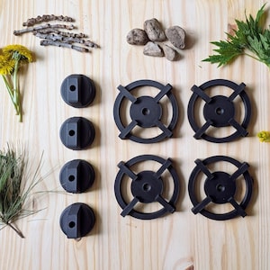 May include: Four black circular stove burner grates and four matching black control knobs arranged in a grid. The items are surrounded by natural elements including yellow dandelion flowers, green pine needles, small twigs, grey stones, and green leaves.