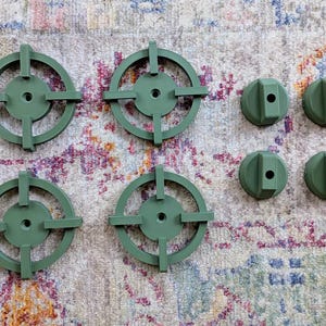 May include: Six olive green, circular objects with a crosshair design and four olive green, rounded, rectangular objects with a hole in the center. The objects are arranged on a patterned, multi-colored surface.
