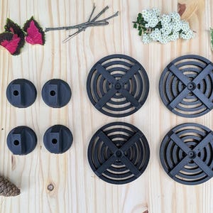 May include: An overhead shot displays several black, circular objects with a cross-shaped design, alongside smaller, rounded black pieces. The items are arranged on a light-colored wooden surface, with natural elements like leaves and a pinecone scattered around.
