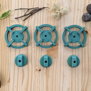 May include: Three teal-colored, circular objects with cross-shaped designs, and three matching smaller, round objects. The items are arranged on a light wood surface, with natural elements like leaves, flowers, and pine cones.