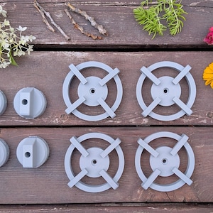 May include: A set of four light gray circular stove burner grates and four matching light gray stove control knobs. The burner grates feature a cross-shaped support structure with a central hole, while the knobs have a cylindrical shape.