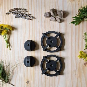 May include: An overhead shot of various natural elements and black metal objects on a light wood surface. The items include green grass, yellow flowers, rocks, twigs, and two black burner grates and knobs.