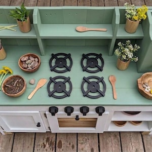 May include: A wooden play kitchen painted in mint green and white. It features a stove with four black burners, a sink, and shelves. Accessories include wooden utensils, bowls with rocks and plants, and flowers. A fun toy for kids.