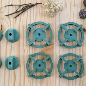 May include: An overhead shot displays teal-colored wooden components, including four circular pieces with cross-shaped designs and four rounded, faceted pieces. The items are arranged on a light-colored wooden surface, accompanied by natural elements like leaves and flowers.