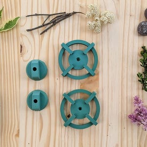 May include: An overhead shot of teal-colored, circular objects with cross-shaped designs, alongside natural elements like pinecones, leaves, and small stones, arranged on a light wooden surface.