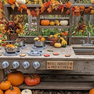 May include: A wooden mud kitchen featuring a stove with four burners, a sink, and a sign reading FALL HARVEST MUD KITCHEN. The structure is decorated with autumn leaves, various pumpkins, and gourds, with metal pots placed on the stove burners.