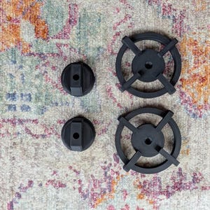May include: Four black stove burner parts are arranged on a patterned rug. Two are circular with cross-shaped supports, and two are smaller, round knobs. The rug has a faded floral design in muted colors.
