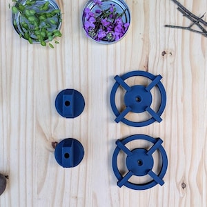 May include: An overhead shot features a light wood surface with various elements. Four small, dark blue, circular objects are arranged with two different designs. Small jars with flowers and greenery are also present, along with a pinecone and sprigs of foliage.