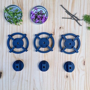 May include: An overhead shot features a collection of items on a light wood surface. There are three navy blue stove burner grates and three matching knobs. Small jars with various herbs and flowers are also present, along with natural elements.