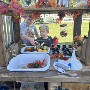 May include: Two young children playing at a wooden outdoor play kitchen. The play area includes a sink, stove, and shelves filled with gourds, leaves, and kitchen tools. A jar of acorns hangs from the top shelf.