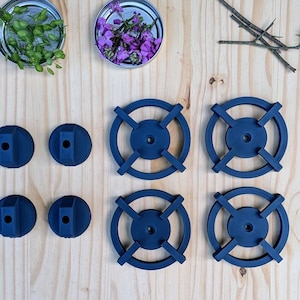 May include: An overhead shot displays a collection of dark blue, circular objects with cross-shaped designs, alongside small jars filled with various plants and greenery. The arrangement is set on a light wooden surface, with additional natural elements like pine needles and a pinecone.