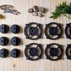 May include: An overhead shot displays black stove burner grates and knobs arranged on a light wood surface, alongside natural elements like twigs, rocks, and greenery. The grates have a circular design.