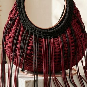 May include: A handcrafted hobo bag with a circular handle. The bag is primarily burgundy with a black top and fringe. The fringe has black and burgundy strands with decorative beads. The bag is labeled "ZALISER | AURA HOBO BAG".