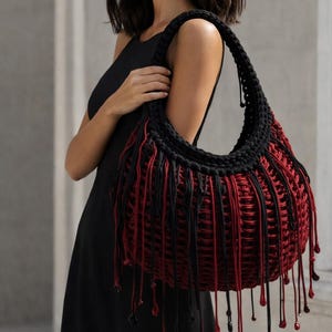 May include: A large hobo-style shoulder bag crafted from dark red and black cord in a woven macrame pattern. The bag features a thick black braided handle and long, dangling fringe accents in alternating red and black colors with small beads.