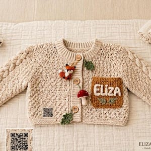 Könnte beinhalten: Beige gehäkelte Baby-Strickjacke mit Zopfmuster an den Ärmeln. Die Jacke hat einen Fuchs- und Pilz-Applikation, eine Tasche mit dem Namen "Eliza" und Holzknöpfe. Der Text "ELIZA'S CLOSET - Handcrafted in Türkiye" ist ebenfalls sichtbar.