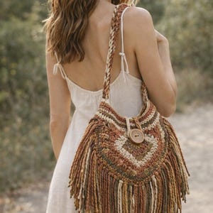 May include: A crochet shoulder bag featuring a geometric pattern in shades of brown, tan, olive green, and cream. The bag has a braided strap, a large wooden button closure at the top center, and long, thick yarn fringe hanging from the bottom edge.