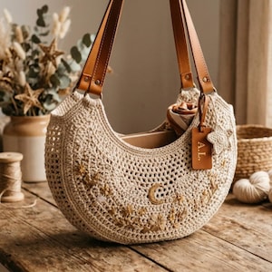 May include: A beige crochet handbag with brown leather straps. The bag features embroidered floral and celestial designs. A brown leather tag with the initials "A.L." hangs from the side. The bag is on a wooden surface.