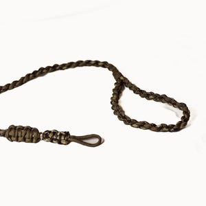 May include: A braided cord in shades of brown and olive green, featuring a loop and a metal clasp. The cord is knotted at one end, and the clasp is a dark, muted metal color. The background is plain white.