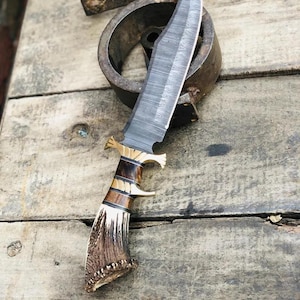 Peut inclure: Un couteau à lame fixe doté d'une lame en acier damas sombre à motifs, d'une garde en laiton et d'un manche composé de segments alternés de bois sombre et de bois de cerf brun texturé, se terminant par un pommeau en bois de cerf naturel.