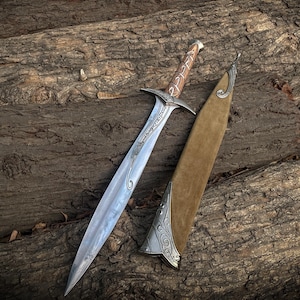 Op de afbeelding: Een tweesnijdend zwaard met een zilverkleurig metalen lemmet met ingewikkelde gravures, een pareerstang en een bruin met leer omwikkeld handvat. Naast het zwaard ligt een bijpassende schede van beige suède met versierde zilveren metalen accenten.