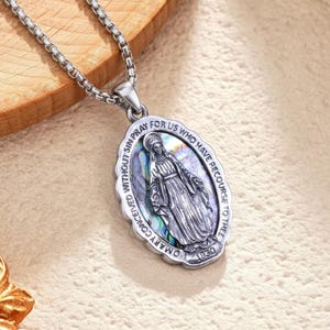 May include: Silver-toned oval pendant necklace featuring an image of a religious figure. The pendant has an iridescent background and is inscribed with text. The necklace chain is a box link style.