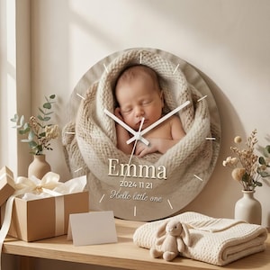 Könnte beinhalten: Eine runde Wanduhr mit einem Baby, das in eine beige Decke gehüllt ist. Das Zifferblatt zeigt den Namen "Emma", das Datum und den Satz "Hello little one". Die Uhrzeiger sind weiß, vor einem beigen Hintergrund.