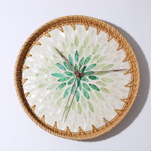 Puede incluir: Reloj de pared redondo con marco de ratán tejido natural. La esfera del reloj presenta un fondo blanco con un diseño floral verde hecho de piezas en forma de hoja. Las manecillas del reloj son de color marrón oscuro.