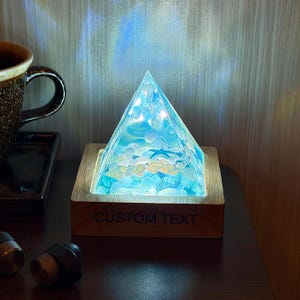 Può includere: Lampada a forma di piramide con un design a conchiglie blu e bianche, posta su una base di legno. La base riporta la scritta "CUSTOM TEXT". La lampada emette una luce soffusa e ambientale, creando un'atmosfera rilassante.