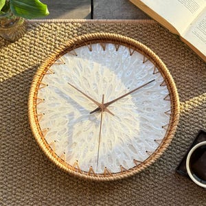 May include: Round wall clock with a woven rattan frame and a white face featuring a leaf-like pattern. The clock hands are brown, and the clock is on a woven mat. The clock measures approximately 12 inches in diameter.