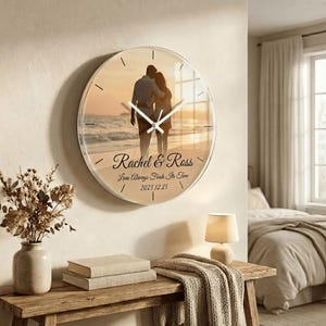 Peut inclure: Une horloge murale ronde et transparente avec une photo d'un couple sur une plage au coucher du soleil. L'horloge a des aiguilles noires et les noms "Rachel & Ross" sont imprimés en script. Le texte "Love Always Finds Its Time" et la date "2025.12.25" sont également imprimés.