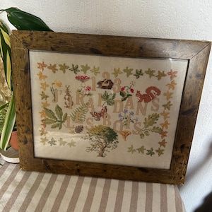 May include: A framed cross-stitch embroidery featuring a border of autumn leaves and a central design of a squirrel, rabbit, butterfly, mushrooms, and a tree. The text "J'AI 3 ANS DANS LES BOIS" is stitched in the centre on a light fabric background.