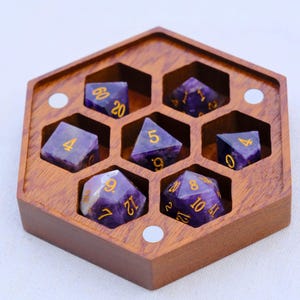 May include: A set of seven purple gemstone dice with gold numbering, presented in a hexagonal wooden box. The dice are arranged in individual compartments within the box. The box is made of a light brown wood.