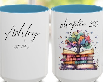 May include: Two white ceramic mugs with blue handles and rims. One mug has the name "Ashley" and "est. 1995" in black script. The other mug has "chapter 30" and a colorful tree design over a stack of books.