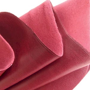 Könnte beinhalten: Ein Stück tiefrotes Leder, das so gefaltet ist, dass sowohl die glatte, bearbeitete Oberseite als auch die weiche, matte und faserige Wildlederunterseite sichtbar sind. Das Material zeigt einen satten, kräftigen Karmesinton mit natürlicher Textur.
