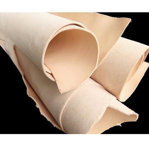 Könnte beinhalten: Ein Stapel hellbeiger, natürlich pflanzlich gegerbter Lederhäute, die zu zylindrischen Formen aufgerollt sind. Das Material zeigt eine glatte, matte Oberflächenstruktur an der Außenseite und ein leicht faseriges, rohes Aussehen an den unebenen Kanten.