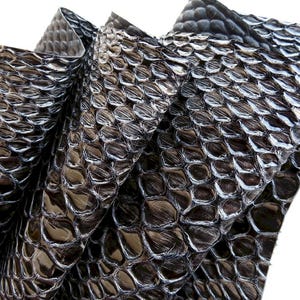 Könnte beinhalten: Ein gefaltetes Stück Schlangenleder in Dunkelgrau und Schwarz mit einer glänzenden, reflektierenden Oberfläche. Das Material weist ein ausgeprägtes, sich wiederholendes Schuppenmuster auf der gesamten Fläche mit tiefen Rillen auf.