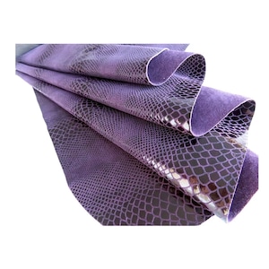 Könnte beinhalten: Ein Stapel lila Kunstlederstoff mit einem Reptilienschuppenmuster. Der Stoff hat eine glänzende Oberfläche auf der gemusterten Seite und eine weiche, Wildleder-ähnliche Textur auf der Rückseite. Die Kanten des Stoffes sind sichtbar.