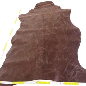 Könnte beinhalten: Ein großes, unregelmäßig geformtes Stück dunkelbraunes Leder mit einer abgenutzten, marmorierten Oberflächenstruktur. Ein gelbes Maßband liegt an der Unterkante und zeigt eine Länge von etwa 100 Zentimetern, ein weiteres liegt an der Seite.