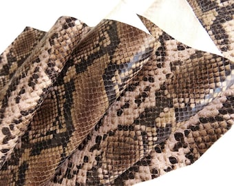 Könnte beinhalten: Ein flexibles Materialstück mit einem realistischen Schlangenhautmuster in Braun-, Beige- und Schwarztönen. Die Oberfläche zeigt eine detaillierte, sich wiederholende Schuppenstruktur mit dunkelbraunen geometrischen Mustern auf einem helleren beigen Hintergrund.