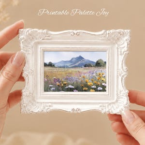 May include: A small, rectangular, ornate white picture frame featuring a landscape painting of a mountain range behind a field of yellow and purple wildflowers. The text "Printable Palette Joy" is printed in a cursive font above the frame.