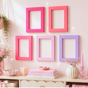 May include: Five rectangular picture frames in shades of hot pink, coral, light pink, and lavender are arranged on a wall above a shelf. The shelf holds a pink glass vase with flowers, a candle, stacked books, and a small pink photo frame.